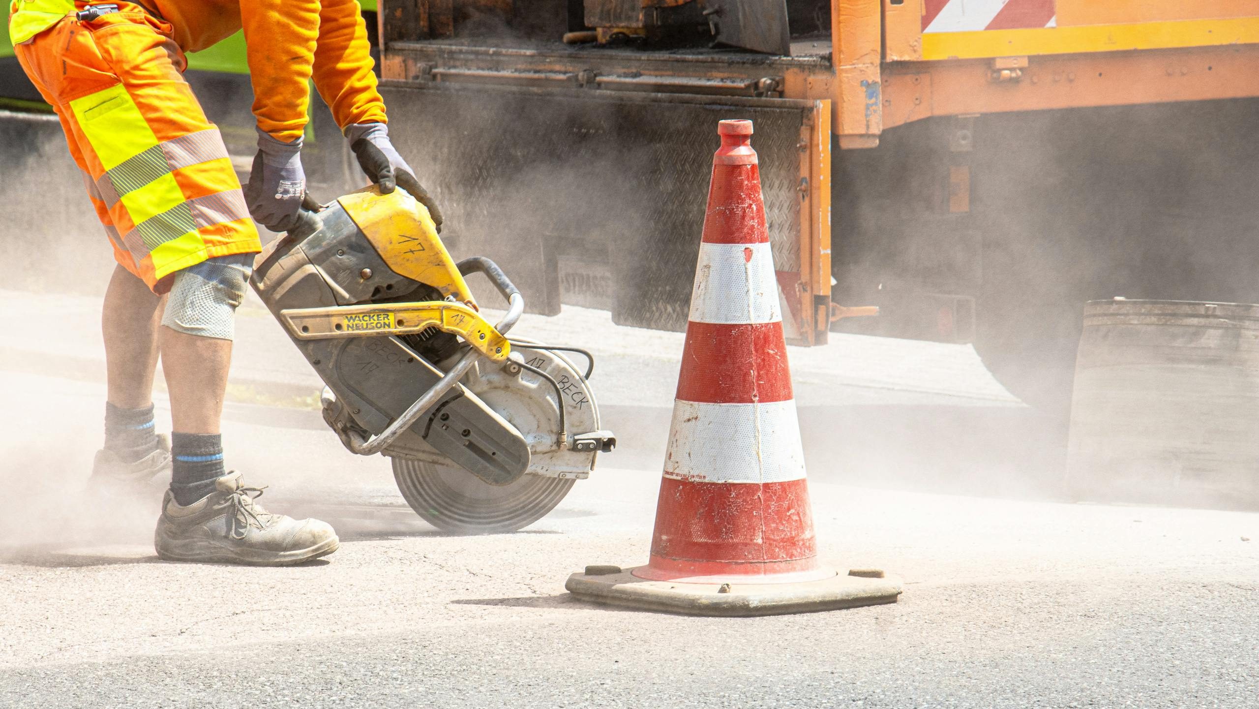A construction worker cutting asphalt with a large saw next to a traffic cone on a street.
